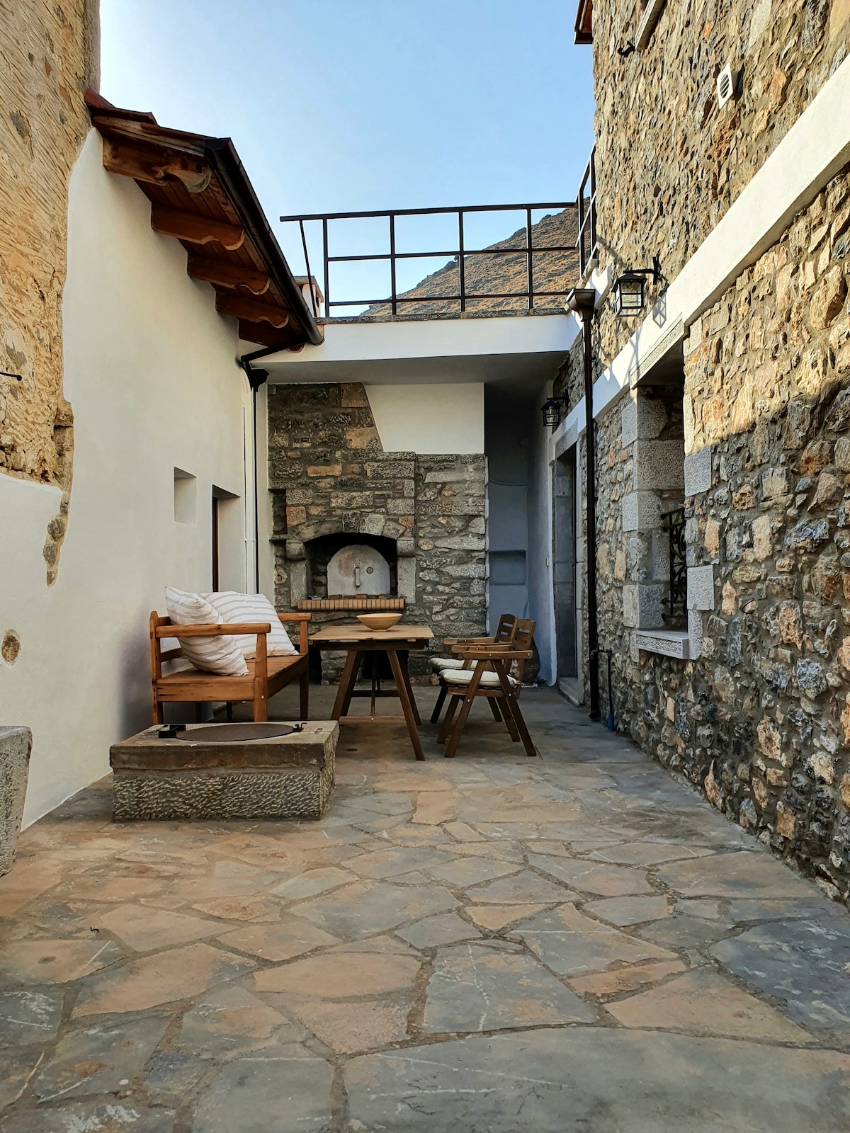 A stone-paved courtyard features a wooden dining table surrounded by chairs. Soft pillows are stacked on a nearby bench, while a stone-built fireplace is visible on one side. The space is bordered by rustic walls and a balcony above, contributing to a cozy outdoor environment.