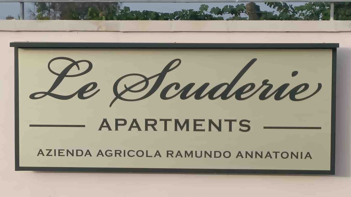 A sign displaying 'Le Scuderie Apartments' is prominently featured, indicating the property’s name. The sign includes the text 'Azienda Agricola Ramundo Annatonia,' highlighting its agricultural connection. The background is softly blurred, with greenery visible, suggesting the location's natural setting.