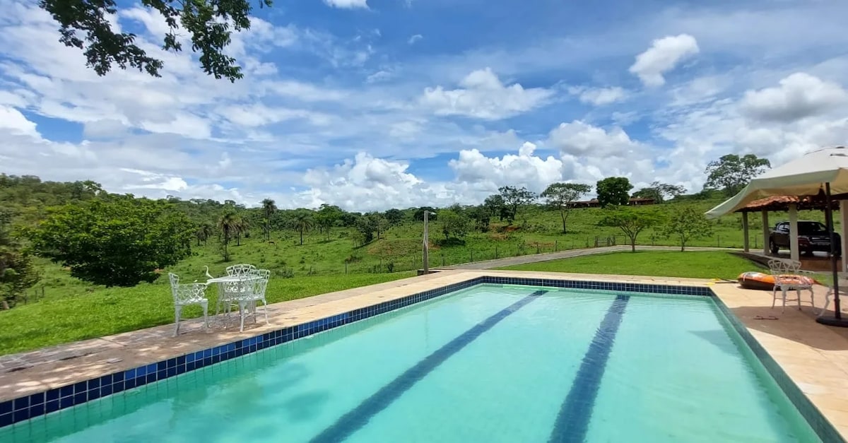 A spacious swimming pool is framed by lush greenery and a clear blue sky. Sunlight reflects off the water's surface, while a white wrought-iron bench is positioned nearby, offering a place to relax and enjoy the tranquil surroundings.