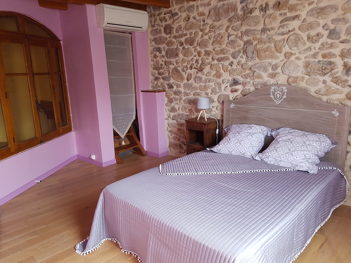 A spacious bedroom features a wooden bed with patterned pillows. Soft purple walls complement a rustic stone wall. Natural light enters through a large arched window with wooden framing, and a small bedside table is positioned next to the bed.