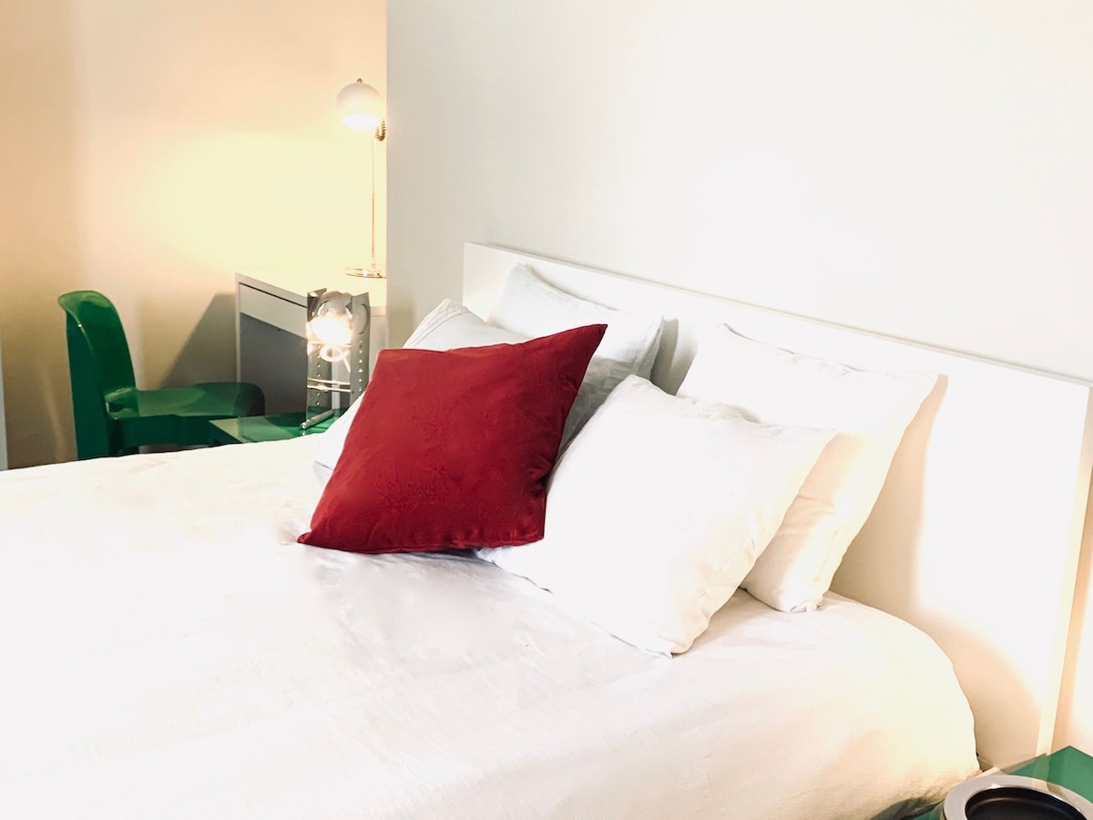 A cozy queen size bed is dressed in crisp white linens, with plush pillows in varying shades. A simple desk is positioned nearby, accompanied by a modern green chair. Soft lighting enhances the serene ambiance of the space.