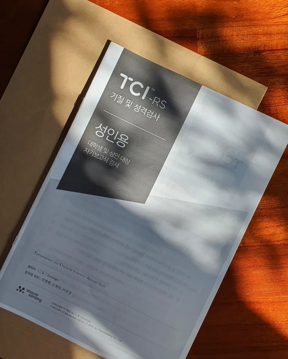 A document is displayed on a wooden surface, with shadows softly falling across it. It features the text 'TCI-RS' prominently at the top, along with other headings in both Korean and English, indicating it is related to personality assessment.