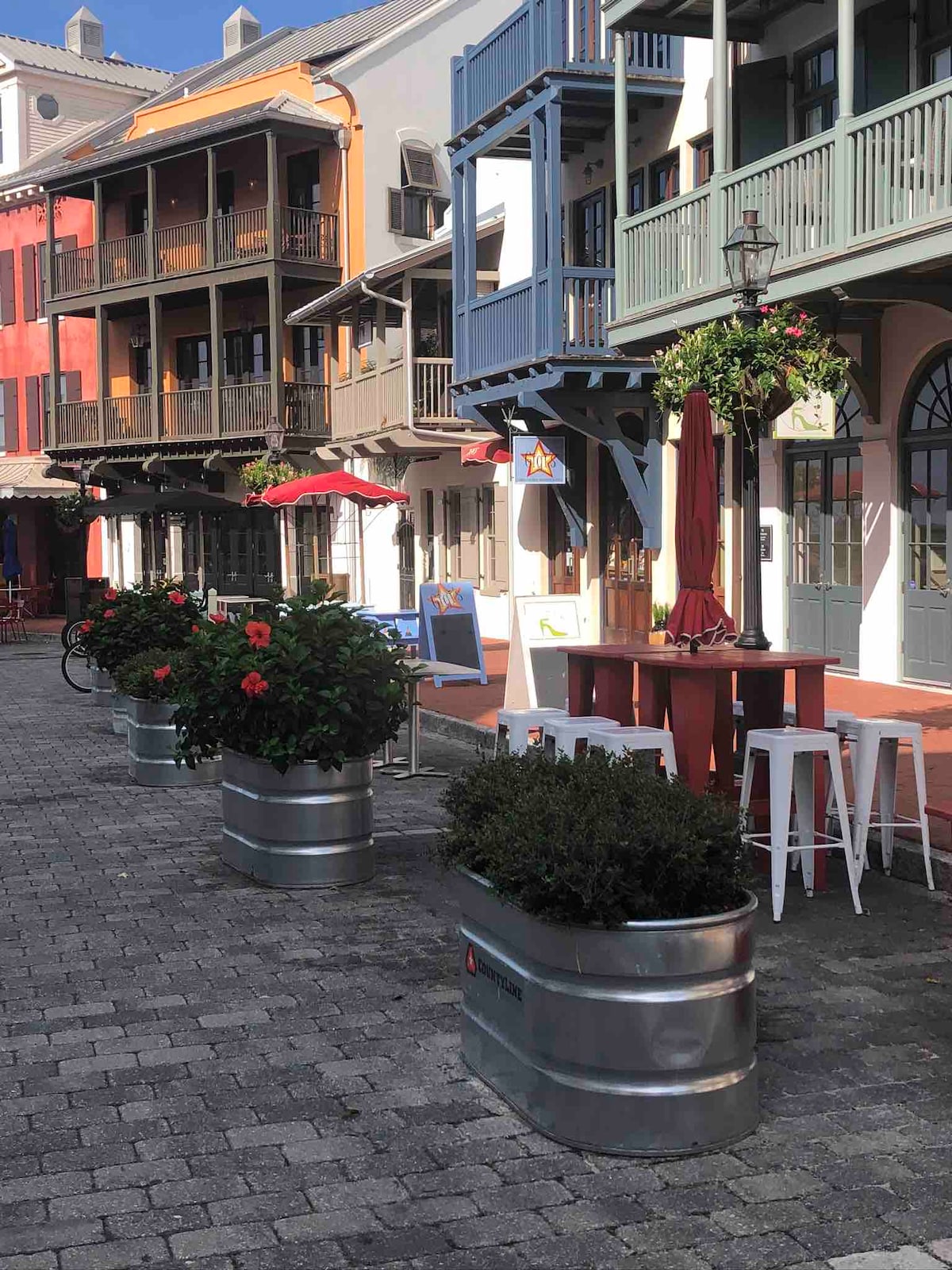 A charming streetscape is depicted, featuring colorful buildings with balconies. Potted plants with vibrant flowers are positioned along the cobblestone pathway, while outdoor seating is provided at a bar table. Umbrellas offer shade, enhancing the casual and inviting atmosphere of the area.