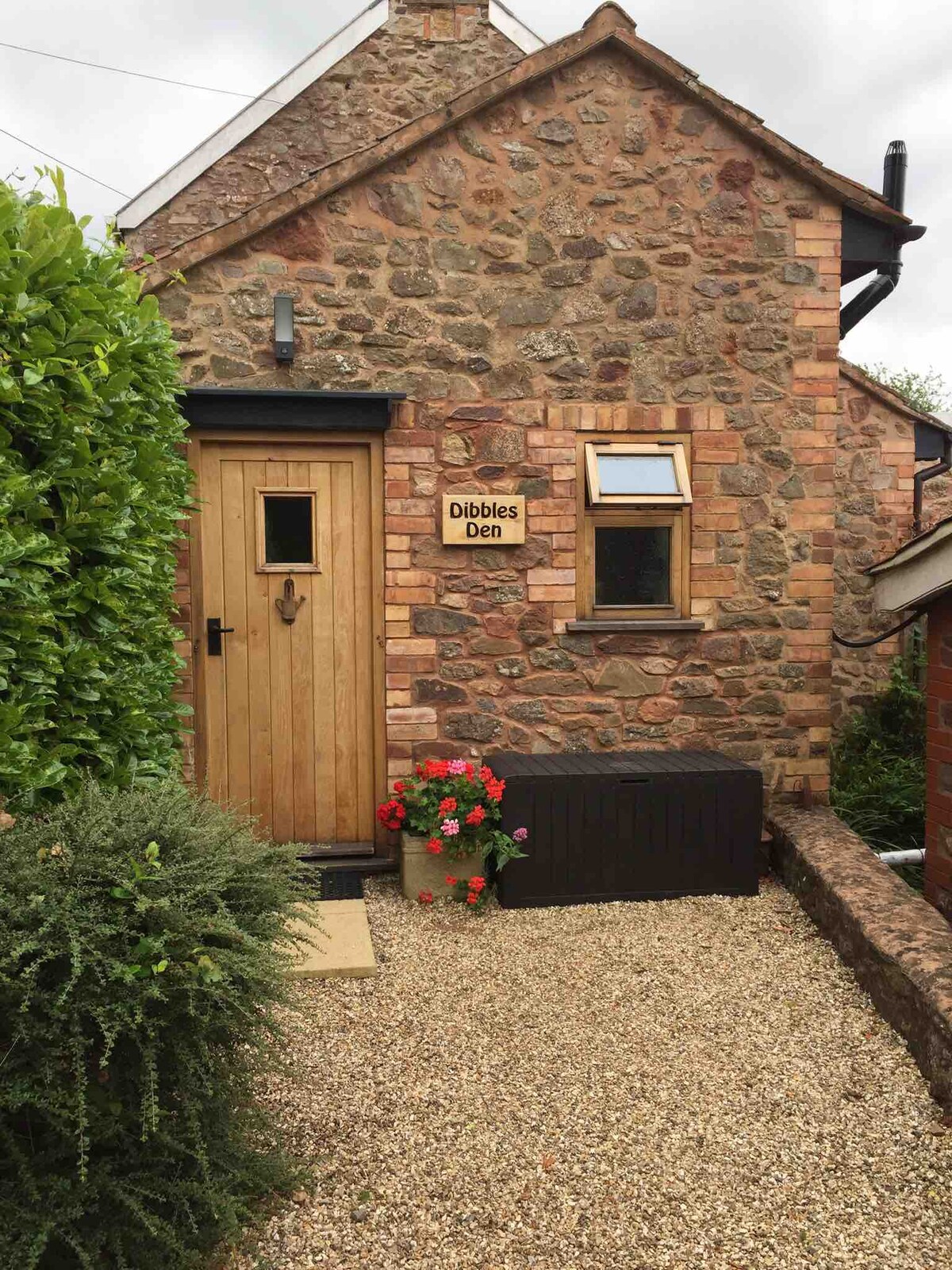 The entrance to the detached annex is marked by a wooden door with a decorative window. A stone exterior is complemented by greenery and vibrant red flowers in a planter. A gravel path leads to the door, providing a welcoming approach.