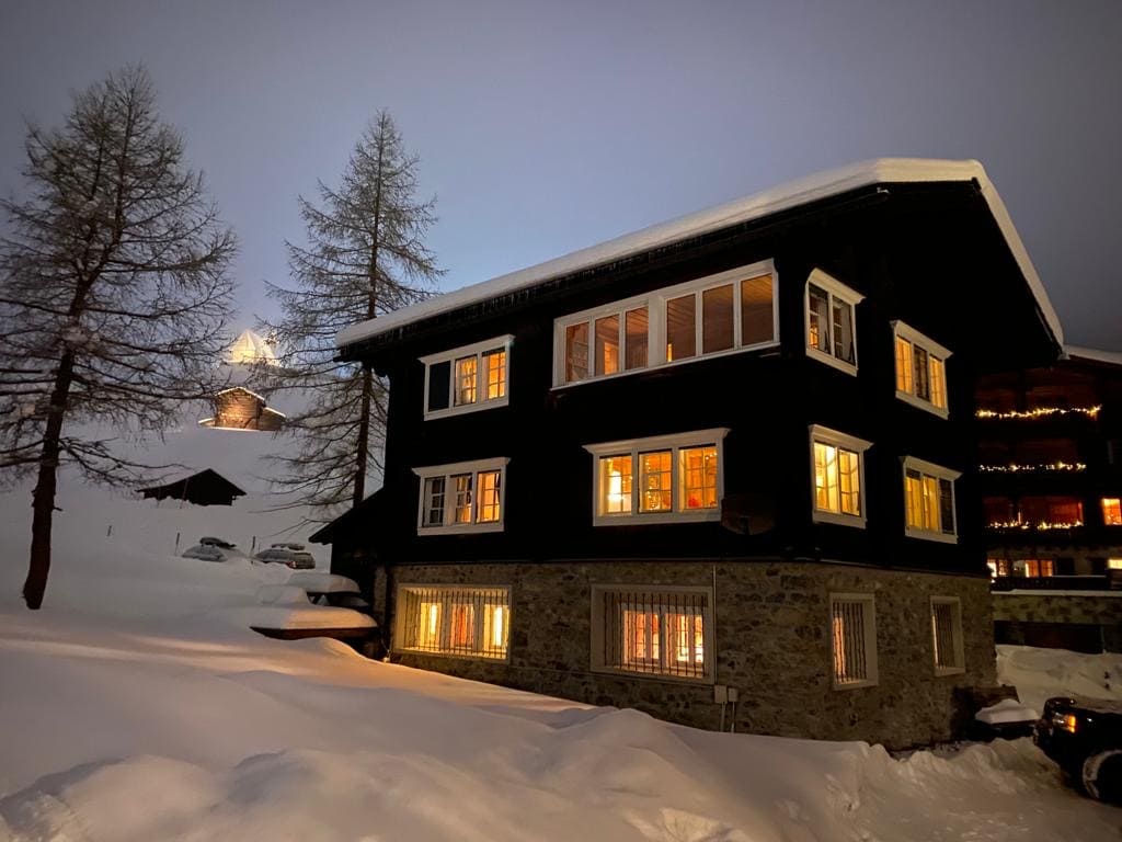 A charming chalet is set against a twilight backdrop, showcasing its dark wooden exterior and large illuminated windows. Snow blankets the ground, while mountains are faintly visible in the distance. The emphasis on the warm glow from inside creates a welcoming atmosphere.