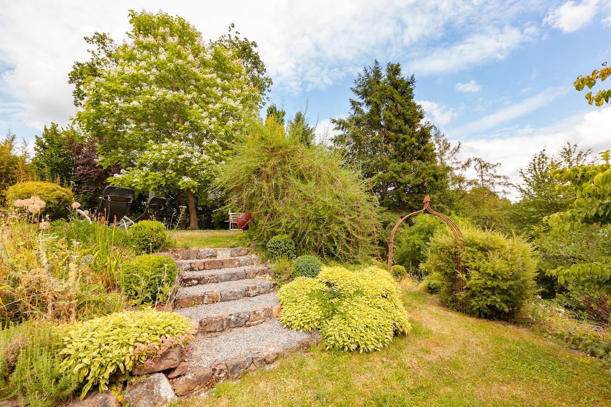 A landscaped garden features a stone pathway leading through greenery and diverse plant life. Large trees provide shade, while various shrubs and flowers create a vibrant and natural environment. The sky is partly cloudy, complementing the calm and serene outdoor space.