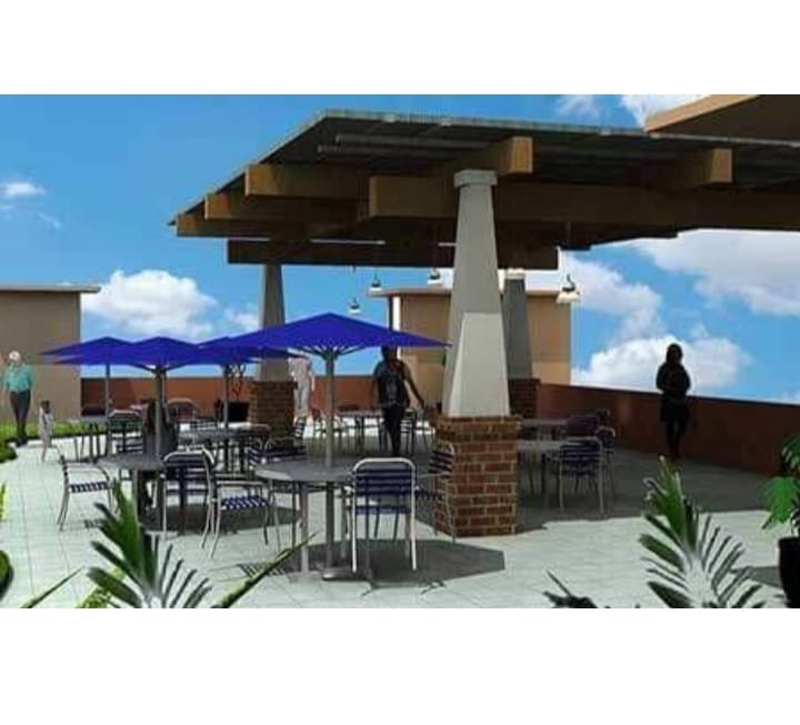 A spacious rooftop terrace is highlighted, featuring several tables and chairs under blue umbrellas. The area is framed by lush greenery, creating a relaxing outdoor space. A clear sky and light clouds are visible, enhancing the overall ambiance.