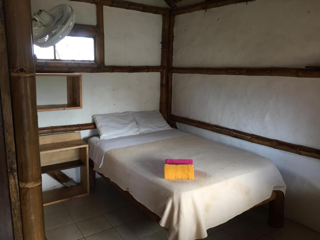 A cozy sleeping area features a comfortable double bed dressed in a neutral-colored blanket. A wooden desk is positioned beside the bed, and a bright towel is neatly placed on top. A window allows natural light to enter, while the walls are finished with a bamboo design.