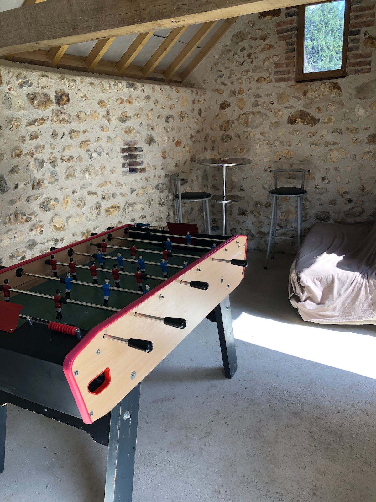 A game room features a foosball table with colorful players waiting for play. Natural light enters through a window, illuminating a small couch and two high stools positioned nearby. The rustic stone walls add character to the inviting space.