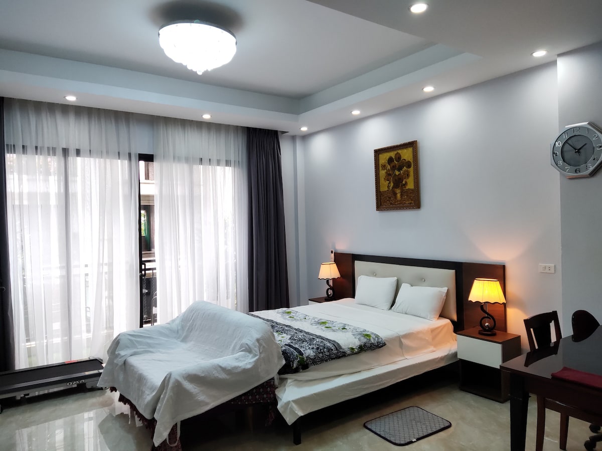 A private studio room features a spacious bed with a light-colored blanket. Soft illumination is provided by bedside lamps. Large windows adorned with sheer curtains allow natural light to fill the space. A small table and a decorative wall art piece complete the room's decor.