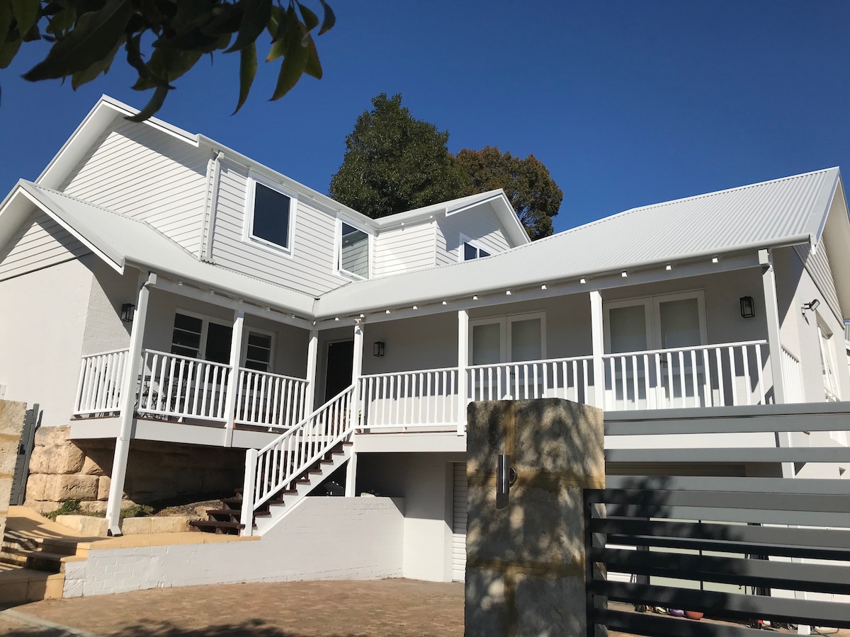 A two-story home featuring a large white porch with railings and steps leading to the entrance. The exterior showcases a mix of white paneling and stone at the base, complemented by a clear blue sky above and greenery from nearby trees.
