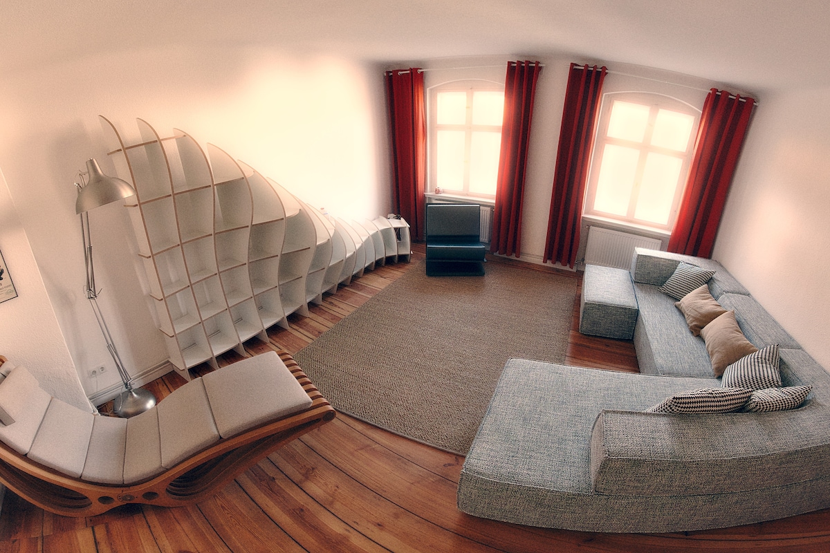 A spacious living area features a large, comfortable sectional sofa and a sleek lounge chair. A unique shelving unit lines one wall, while windows dressed with red curtains allow natural light to filter in. A neutral area rug anchors the space, complementing the wooden floor.
