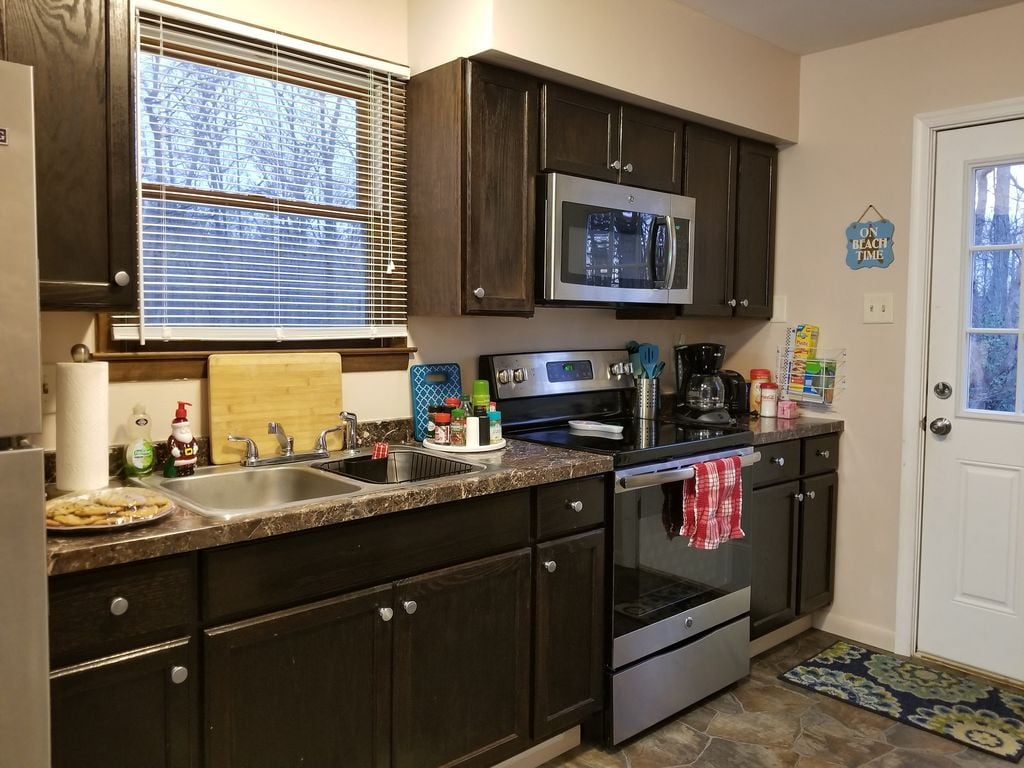 A well-equipped kitchen features dark cabinetry and modern appliances, including a microwave and stove. A double sink is positioned beside a window that overlooks a wooded area. Various kitchen supplies and condiments are neatly arranged on the countertop, contributing to a functional cooking space.