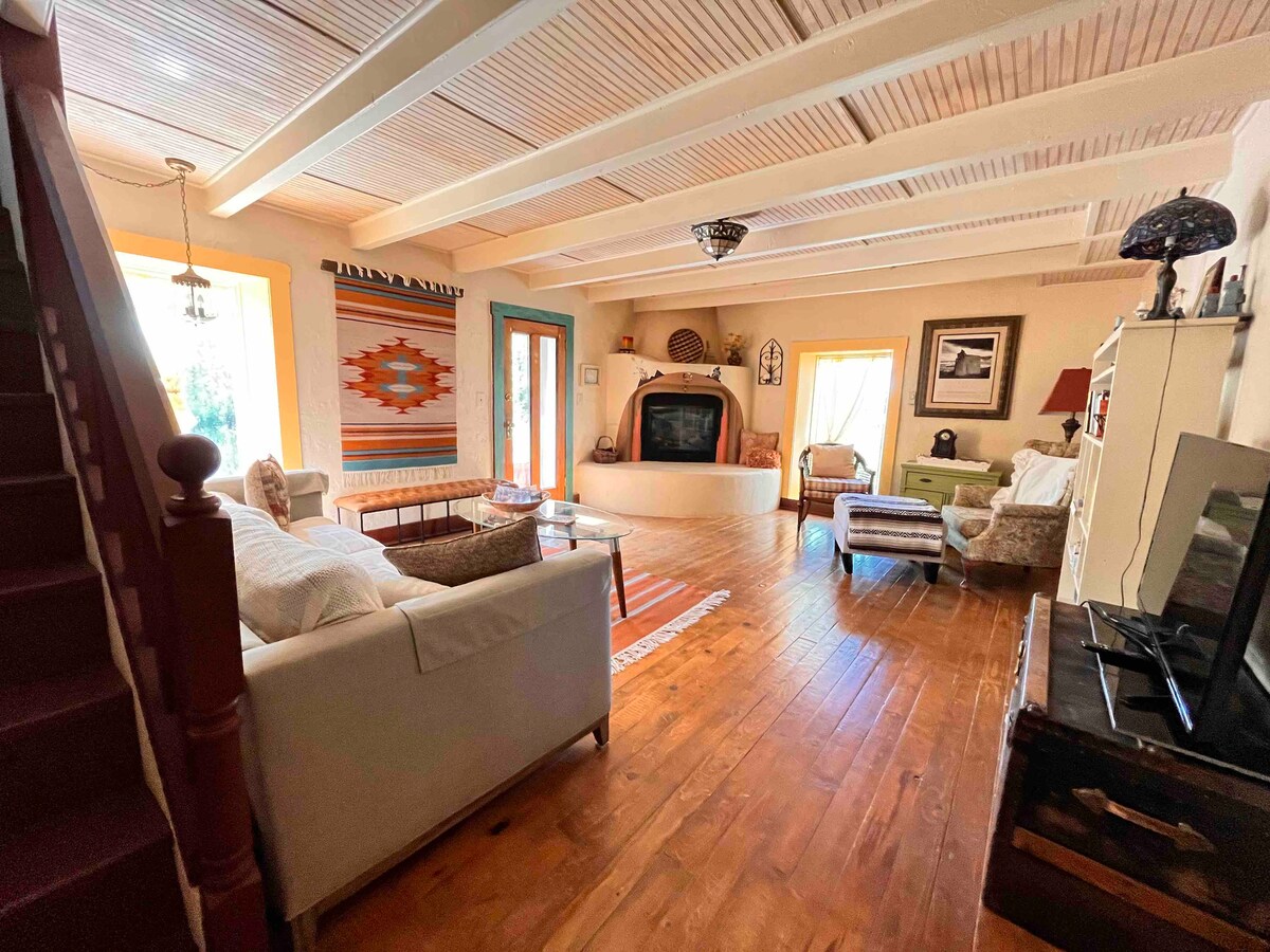 A spacious living area showcases wooden flooring and exposed beams. A cozy seating arrangement is highlighted by a central fireplace and large windows. Colorful textiles and decorative pieces adorn the walls, enhancing the room's warm and inviting character.