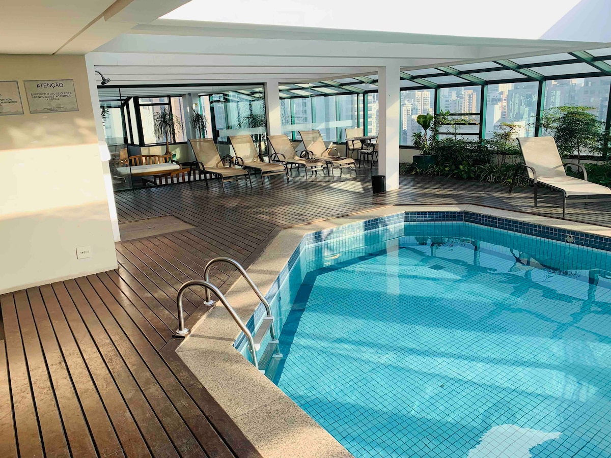 A bright indoor pool area is surrounded by large glass windows, allowing natural light to fill the space. Lounge chairs are arranged along the wooden deck, offering areas for relaxation. The pool features a textured surface and a ladder for easy access.