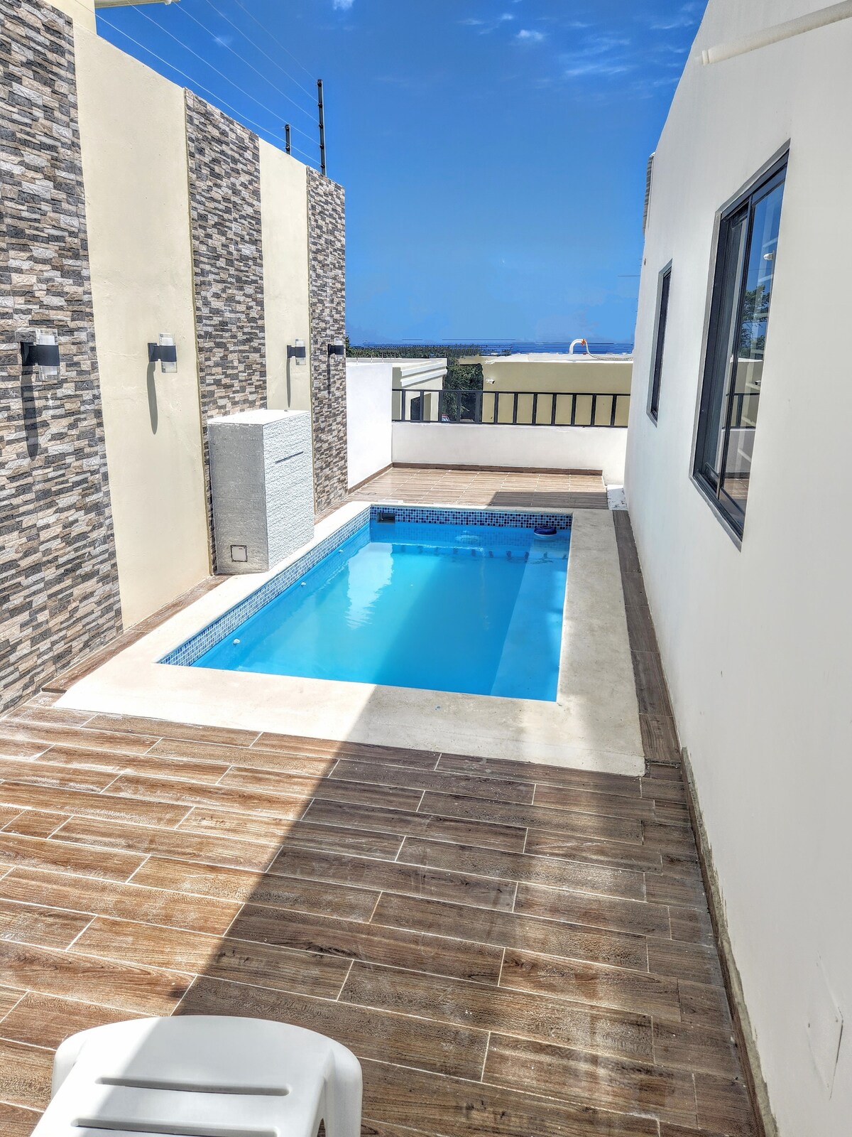 A private pool is framed by neutral-toned walls and stylish stone accents, reflecting the clear blue sky above. An inviting wooden deck surrounds the pool, providing a tranquil space for relaxation.
