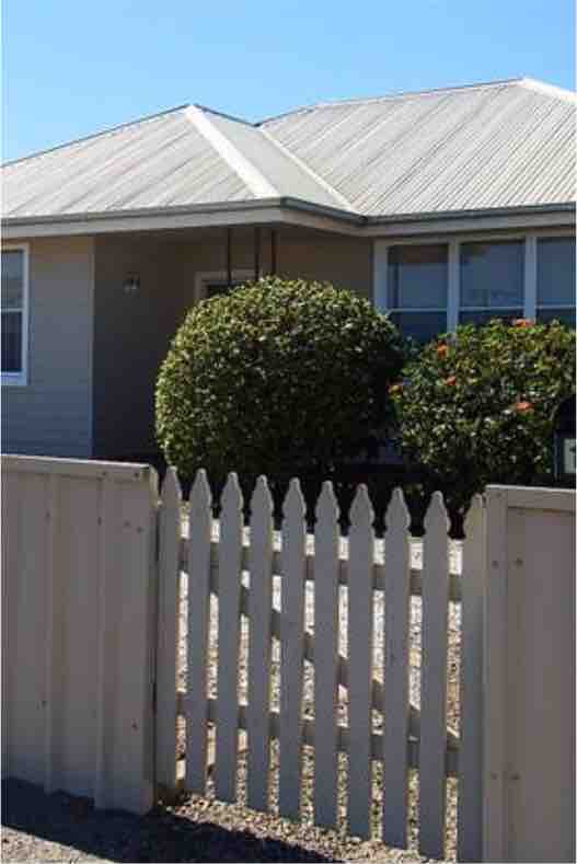 A modest, single-story house is viewed from outside, featuring a grey facade and a distinct gabled roof. The front yard is neatly presented with vibrant greenery, including shaped shrubs, and is enclosed by a pale picket fence.