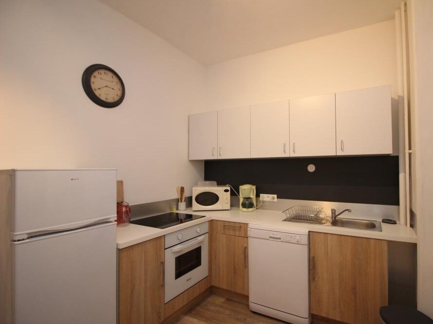 The kitchen area features modern cabinetry in light wood and white finishes. A refrigerator, oven, dishwasher, and microwave are included. A kettle and coffee maker are visible, along with essential cooking utensils. A clock adorns the wall, providing a convenient reference.