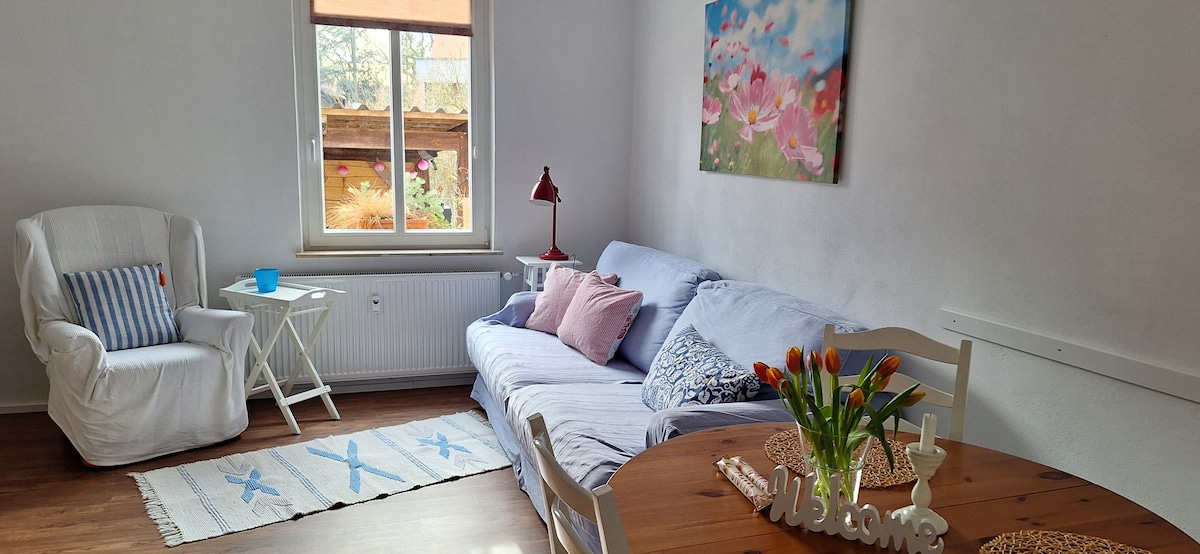 A bright living area features a comfortable sofa adorned with pastel cushions, complemented by a white armchair. A small table holds a blue glass and a decorative piece, while a colorful floral painting adds a touch of vibrancy to the wall. Natural light filters through the window.