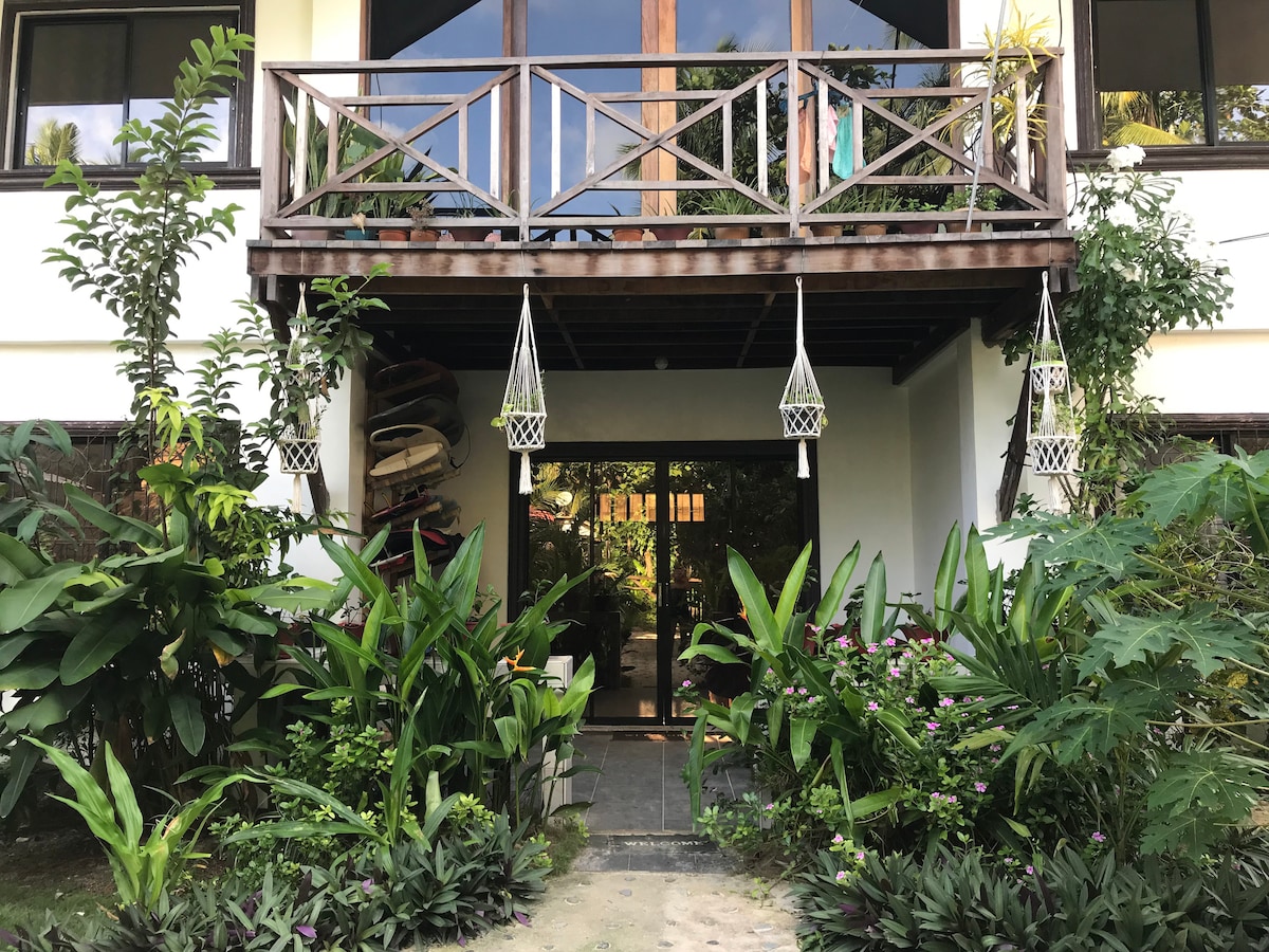 The entrance is framed by lush greenery and blooming plants. A wooden balcony with a railing is visible above, adorned with hanging decorations. Surfboards are stored against the side, while natural stone pathways lead to an inviting front door.
