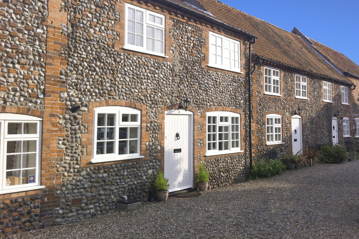 The exterior of the cottage is constructed from traditional Norfolk flint and brick, showcasing a textured stone facade. Multiple windows, framed in white, allow natural light to enter. A central front door, bordered by potted plants, provides a welcoming entry to the property.