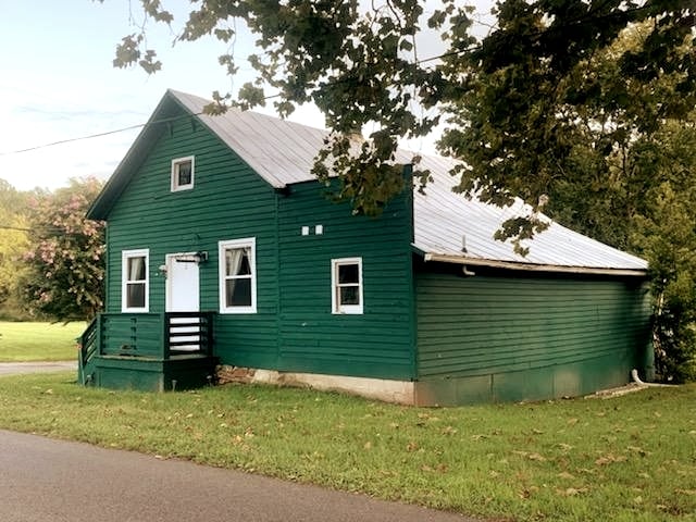 The exterior of the rustic green cottage is shown, featuring a metal roof and a welcoming porch. Surrounding trees provide a natural setting, and a grassy area leads to the driveway. Multiple windows allow natural light into the home.