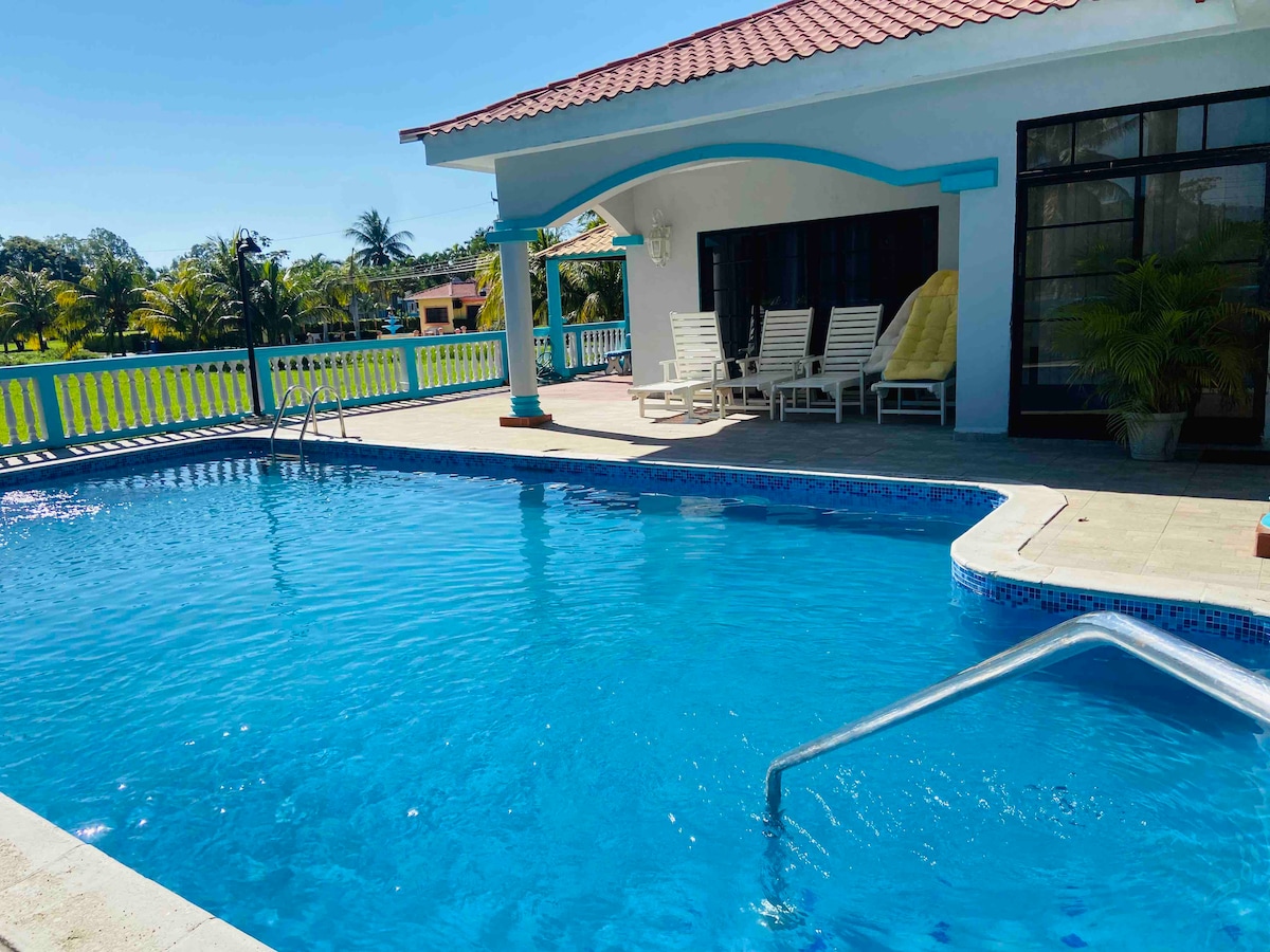 A large swimming pool is visible, surrounded by a spacious patio area. Sun loungers are arranged against the wall under a sheltered porch. Lush greenery and palm trees frame the setting, creating a bright and inviting outdoor space.