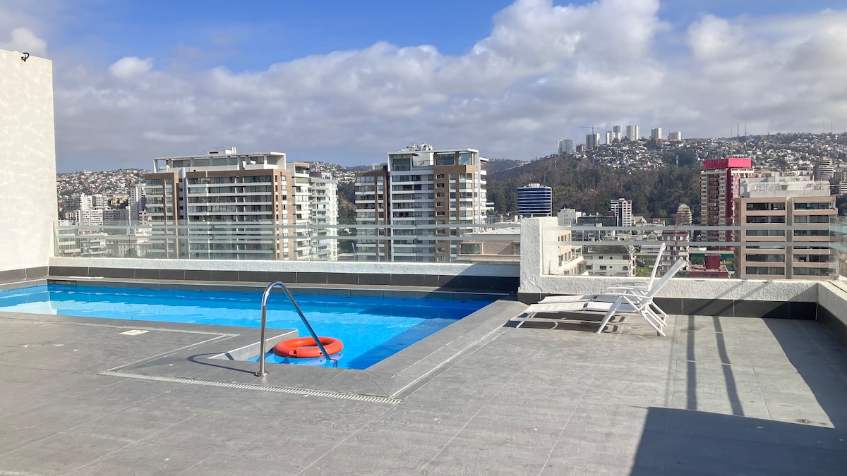 A rooftop pool area offers a bright swimming pool with a safety ring, surrounded by a panoramic cityscape. Lounge chairs are positioned for relaxation, while the clear sky enhances the spacious outdoor setting. The contemporary design reflects a blend of urban living and leisure.