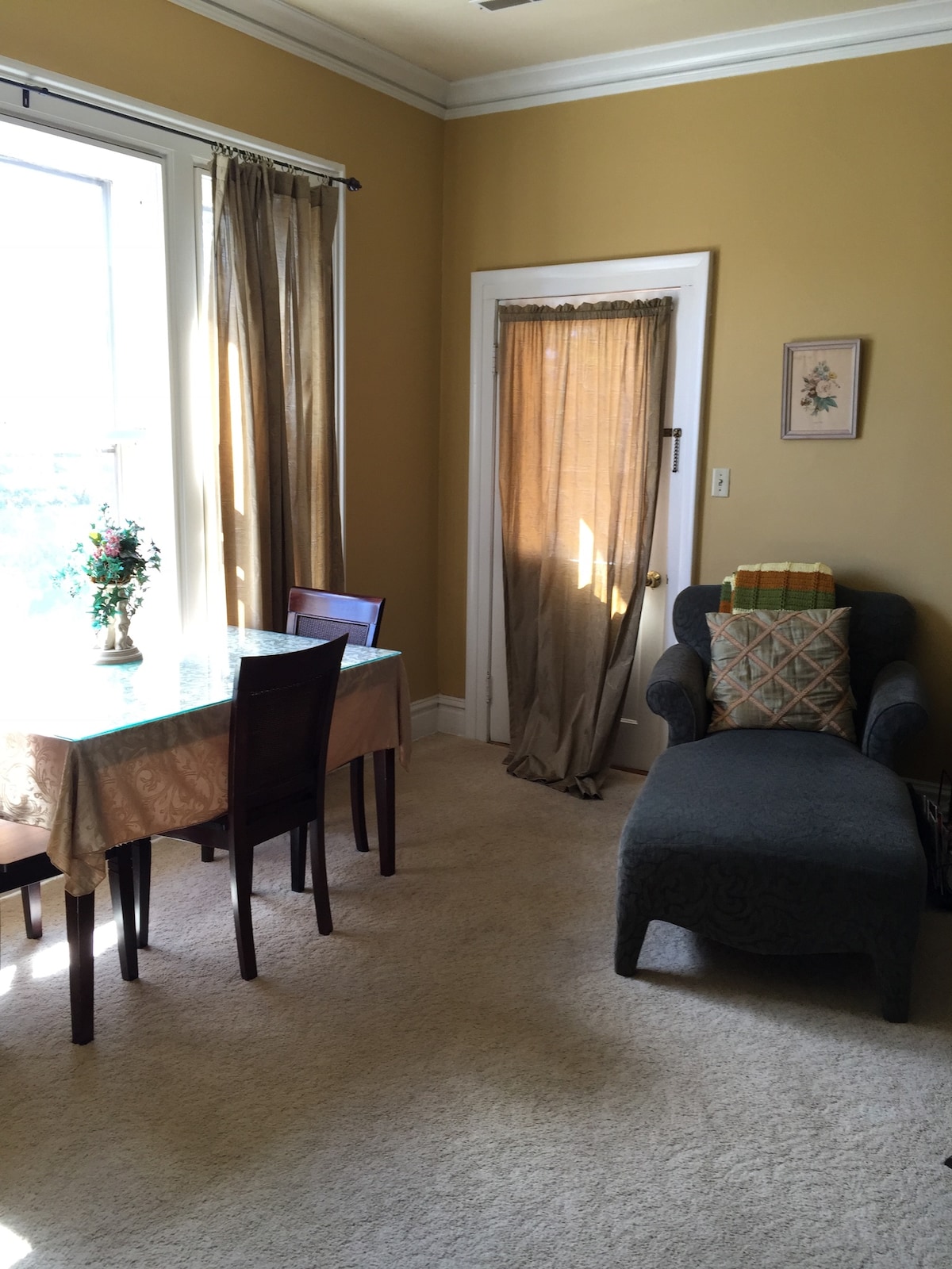 A sunlit corner features a small dining table with four chairs, accompanied by a floral centerpiece. A cozy armchair with a patterned cushion sits nearby, while sheer curtains allow natural light to filter in. A door leads to an adjacent space, framed by soft drapery.
