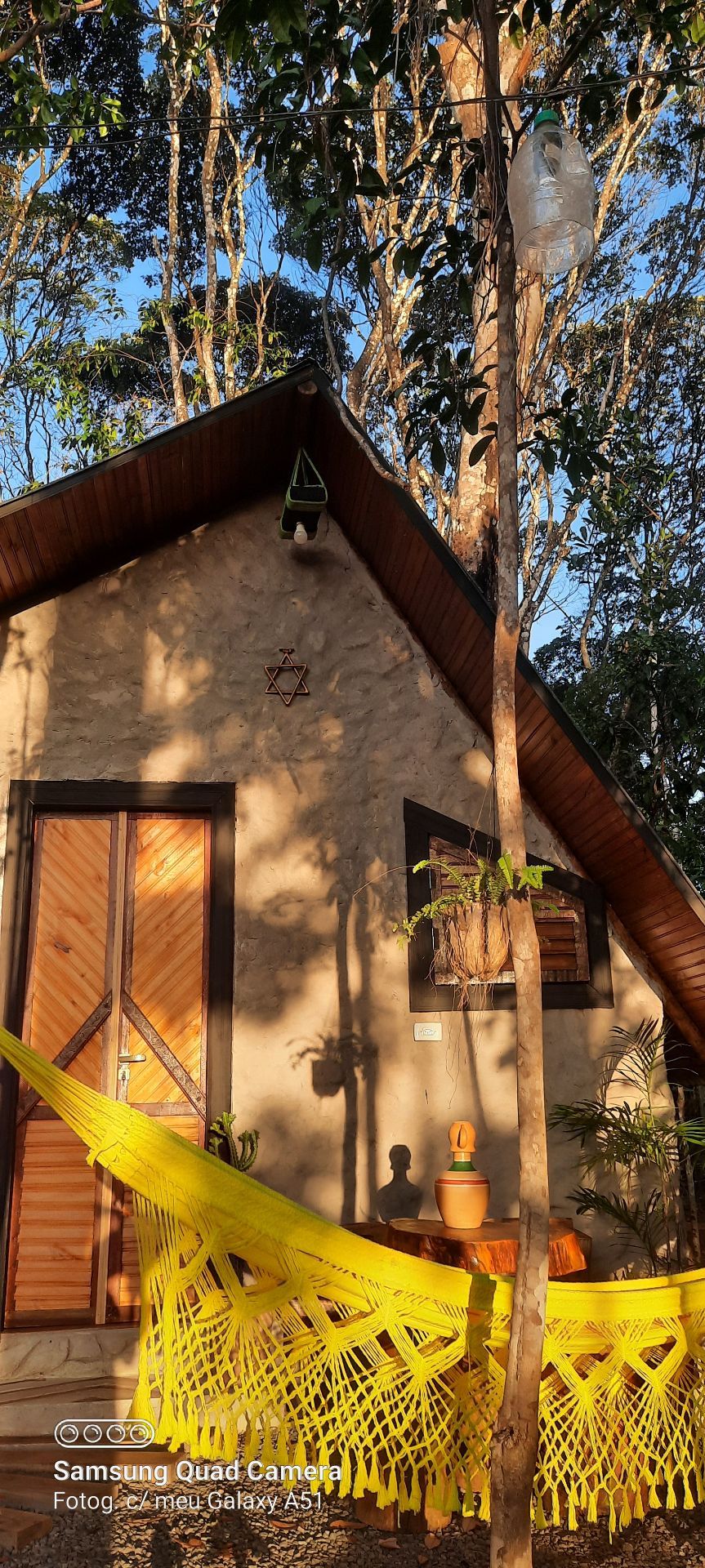 An entrance to a cozy cabin is framed by lush greenery. The structure features a sloped roof and wooden doors, with a colorful yellow hammock hanging nearby. A small potted plant is positioned at the base of the cabin, reflecting a tranquil outdoor space.