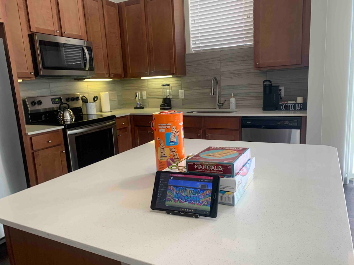 A modern kitchen is shown with wooden cabinetry and stainless steel appliances. A central white countertop is highlighted, featuring a colorful game box, a tablet, and a tin container. Natural light streams in through the window, enhancing the inviting atmosphere.
