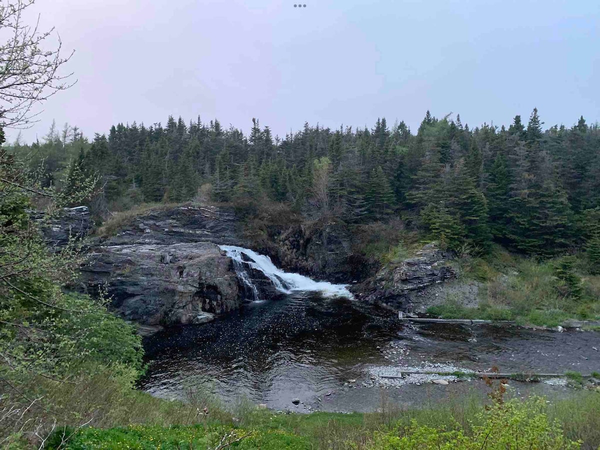 A tranquil scene of a waterfall cascading over rocks into a calm body of water, surrounded by lush trees. The greenery is interspersed with rocky formations, creating a natural setting that reflects the serenity of the area.