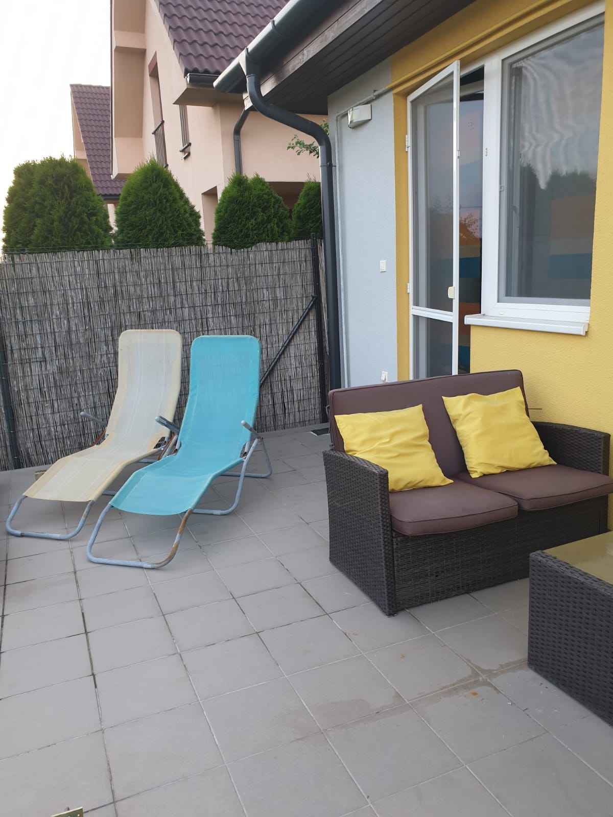 A terrace area features two reclining beach chairs alongside a cozy seating arrangement with a wicker sofa and yellow cushions. A small table is placed in front of the seating. A privacy screen and lush greenery can be seen in the background.