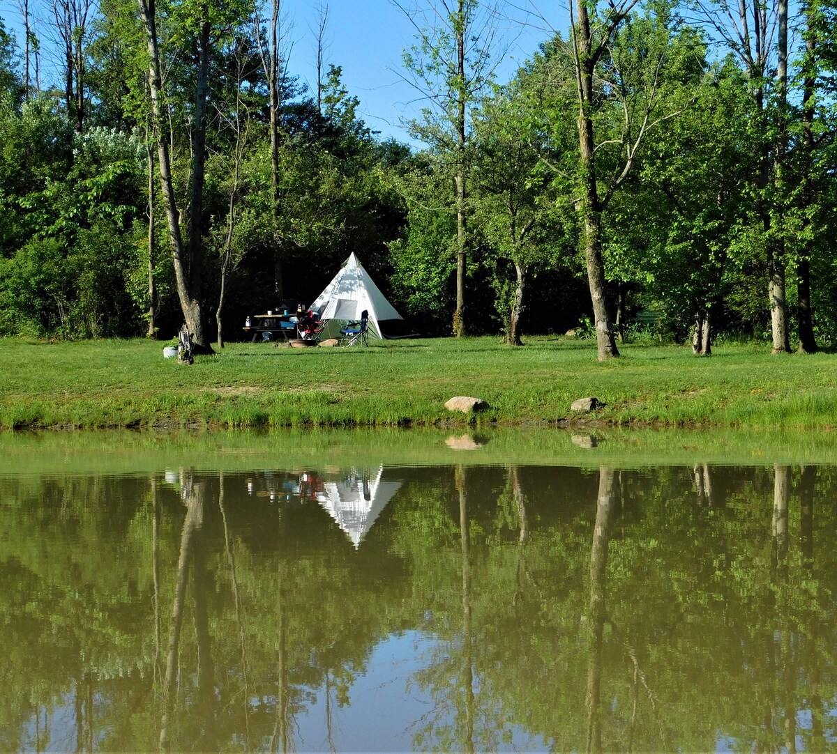 A traditional tent stands on a grassy campsite, enveloped by trees and greenery. A picnic table is positioned nearby, and reflections of the tent and trees can be seen in the calm water of a small pond in the foreground.