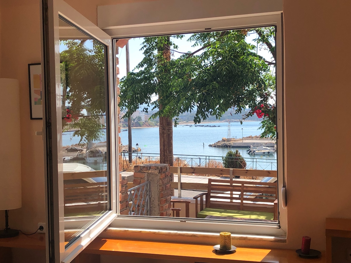 A window opens to a view of the waterfront, featuring boats docked in the harbor. Green trees frame the tranquil scene, while seating areas are visible along the shore. Natural light enhances the inviting indoor space.
