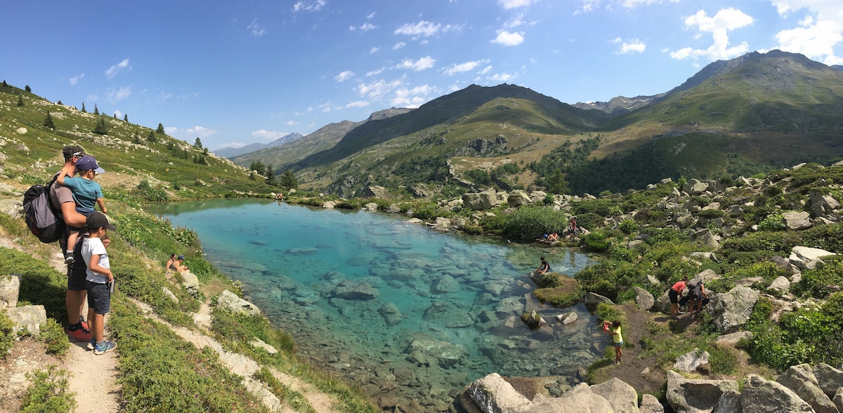 A panoramic view captures a serene alpine lake surrounded by lush greenery and rocky terrain. People can be seen enjoying the water and sunbathing along the shore. Mountains rise in the background under a partly cloudy sky, creating a tranquil natural setting.