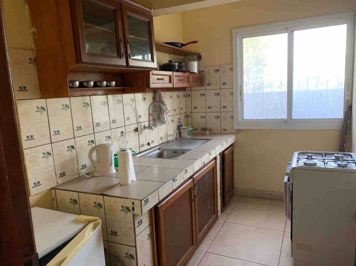 The kitchen features wooden cabinets and a tiled backsplash. A double-basin sink is situated beneath a large window, allowing natural light to enter the space. An oven and refrigerator are present, along with various kitchen essentials on the countertops.