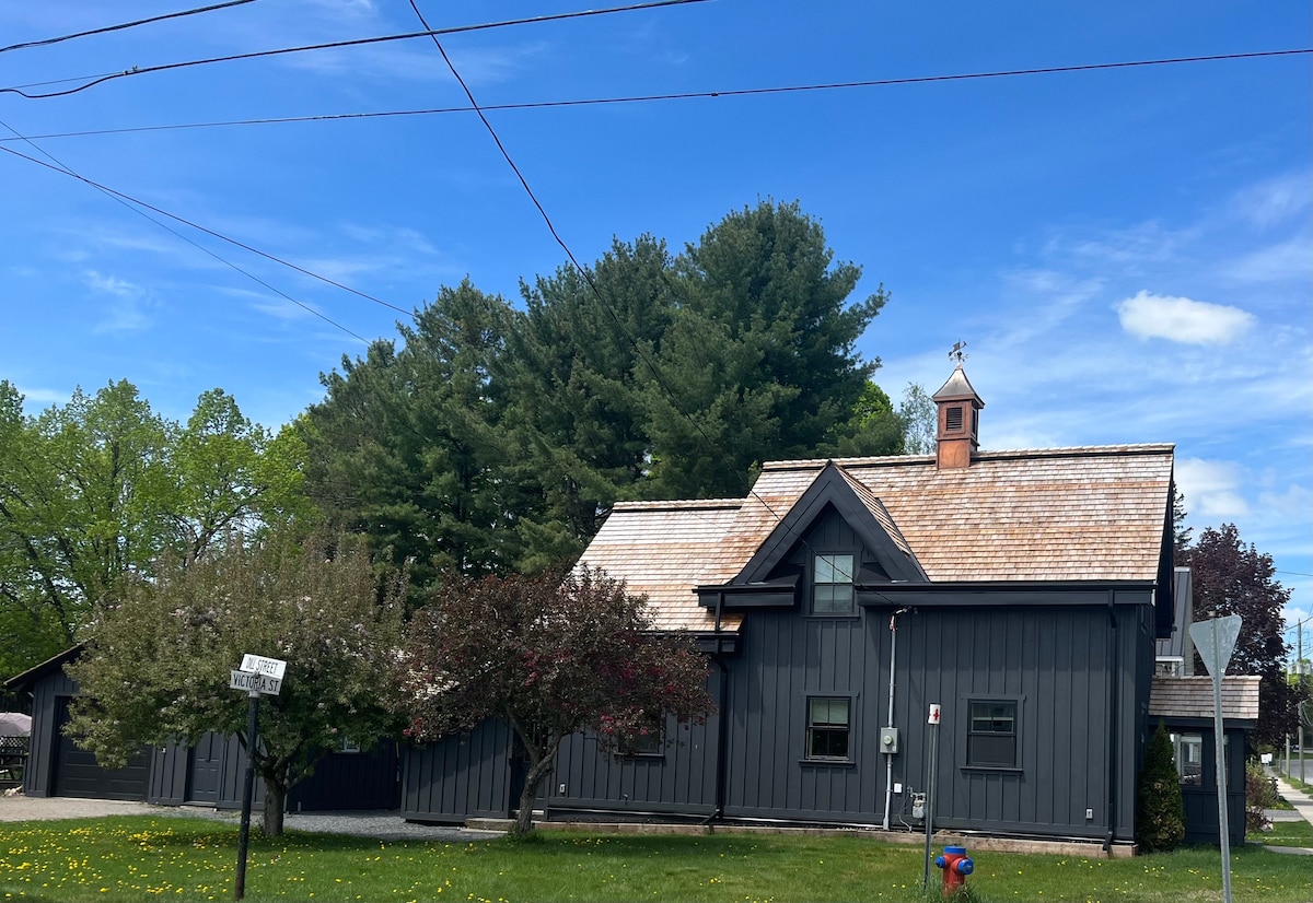 The exterior of a modern black timber house is framed by lush green trees and blooming landscaping. A pitched roof features a decorative cupola, while windows are adorned with dark trim. The clear blue sky complements the welcoming facade, enhancing the property’s charm.