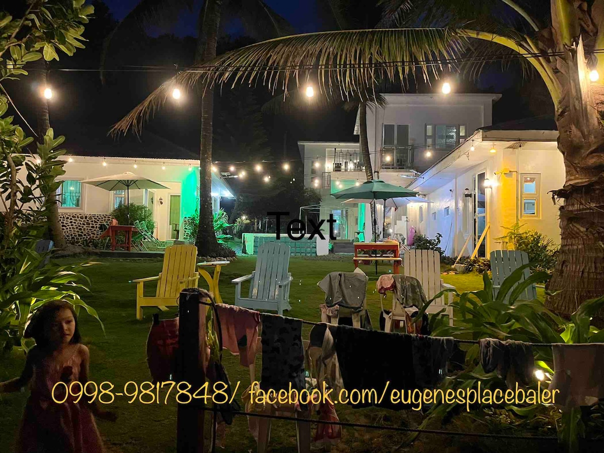 A vibrant outdoor space is illuminated by string lights at night. Colorful villas with varying wall colors, including yellow and green, surround a grassy area. Lawn chairs are arranged for seating, and clotheslines with hanging garments add a homey touch.
