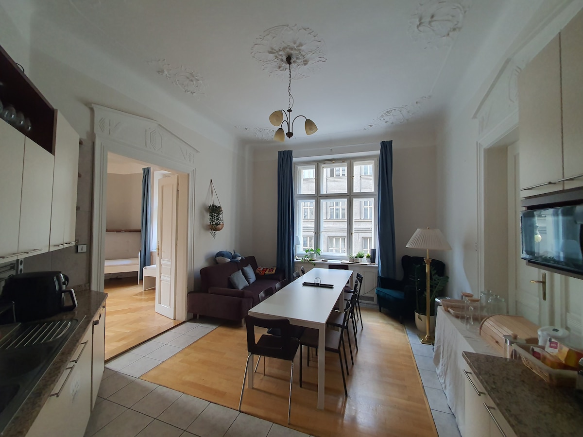 A spacious kitchen and dining area is shown, featuring a long table with seating for multiple guests. Natural light filters in through a large window, accentuating the light-colored cabinets and countertops. Nearby, a cozy seating area with a plush sofa and lamp is visible.