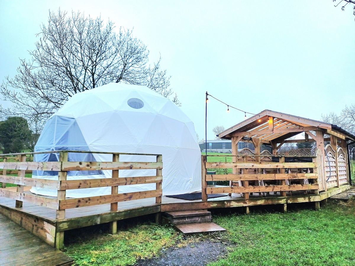 A geodesic dome is surrounded by a wooden deck, featuring a canopy and outdoor seating area. String lights are strung overhead, enhancing the cozy ambiance. A grassy area surrounds the structure, with bare trees in the background, indicative of a tranquil rural setting.