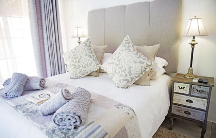 A neatly arranged sleeping area features a spacious bed with a light-colored headboard, adorned with patterned throw pillows. Coordinating blankets are neatly placed at the foot of the bed. Soft lighting is provided by bedside lamps, enhancing the calm ambiance of the space.