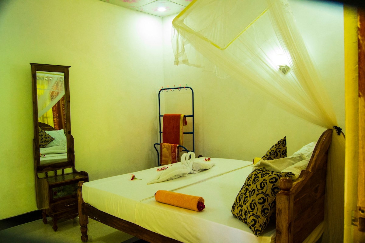 A cozy sleeping area features a wooden bed adorned with decorative pillows. A large mirror stands beside the bed, and a clothing rack is positioned nearby. Soft lighting creates a calm ambiance, complemented by a mosquito net draped over the bed.