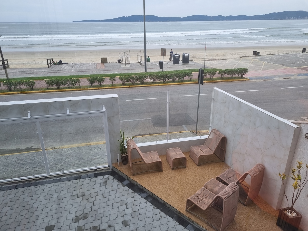 An outdoor seating area features contemporary lounge chairs positioned on a textured surface, with views of the beach and ocean. A low glass barrier separates the space from the street, while a few potted plants add subtle greenery to the scene.