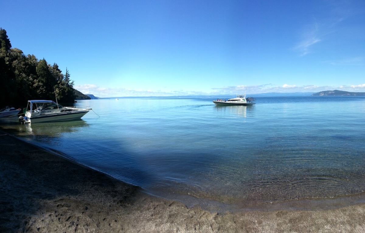 A serene view of Lake Taupo is presented, with calm waters reflecting the clear blue sky. A boat is seen approaching the shore, while trees border the left side of the image, enhancing the natural setting.