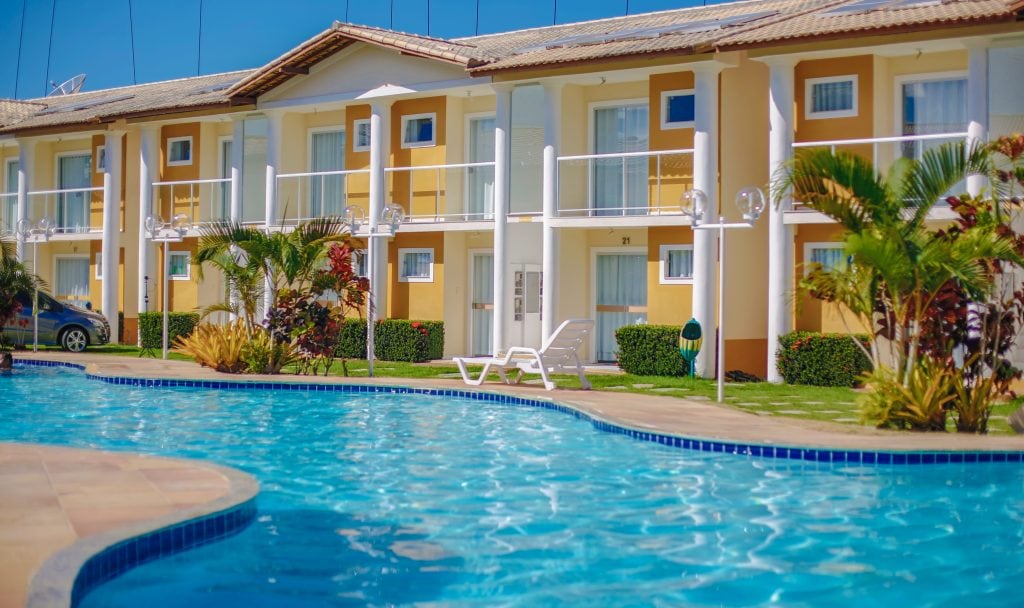 The exterior of a two-story residential building is seen, featuring a refreshing pool in the foreground. Lush greenery and colorful plants surround the area, with spacious balconies visible on each unit. Sun loungers are positioned by the pool, creating a peaceful environment.