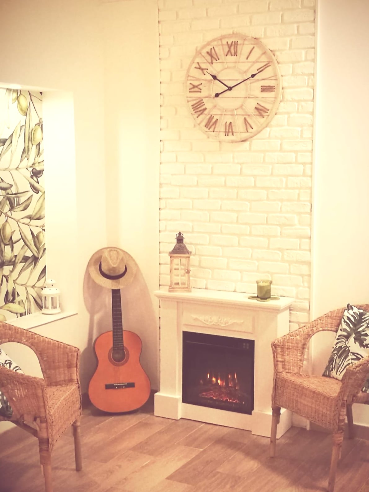 A cozy corner features a rustic electric fireplace surrounded by two wicker chairs. A wooden clock with Roman numerals is mounted above, while a guitar leans against the wall. A decorative lantern and a green drink sit on the mantel, enhancing the relaxing atmosphere.