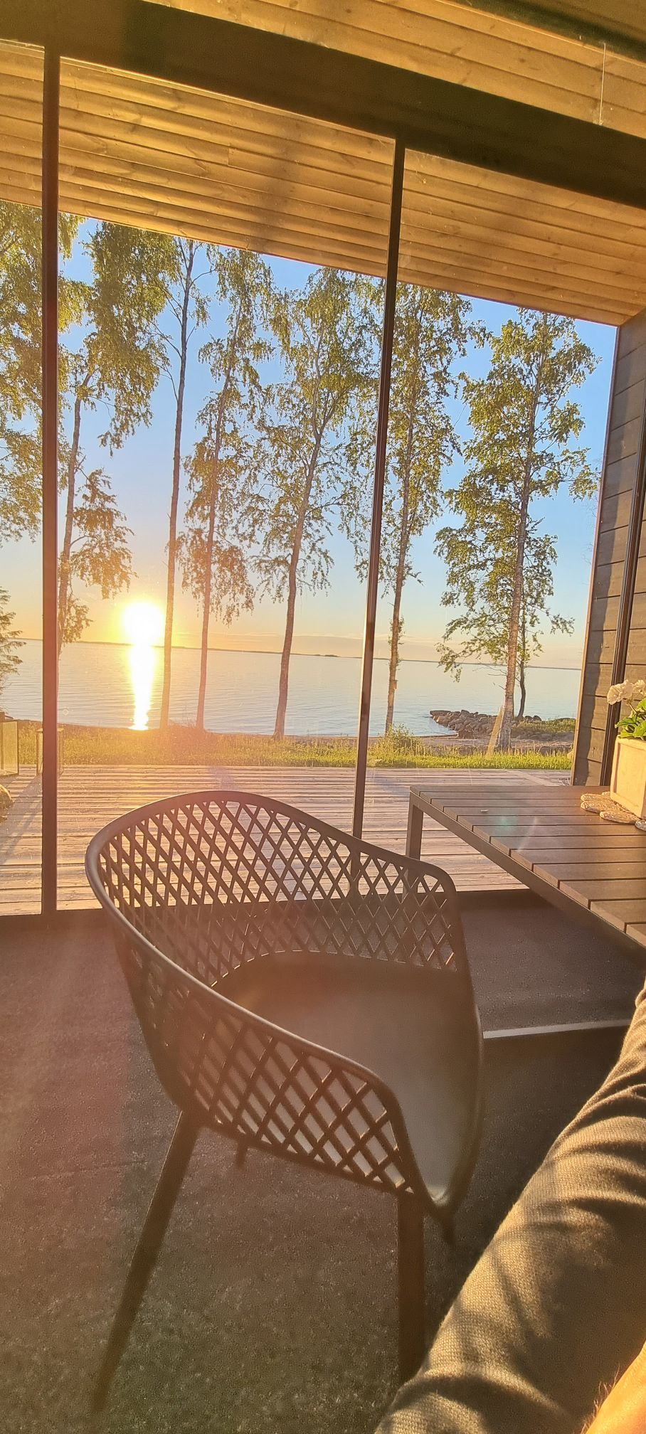 A glass wall offers a stunning view of the sun setting over the water, with reflections shimmering on the surface. A black woven chair is positioned on a deck, complemented by a backdrop of trees and a serene lakeside environment.