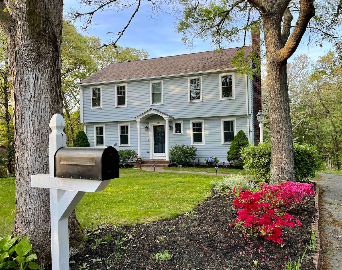 A two-story home with light blue siding and white trim is set amid lush greenery and flowering shrubs. A mailbox stands near the entrance, framed by well-maintained grass and colorful flower beds, enhancing the home's welcoming curb appeal.