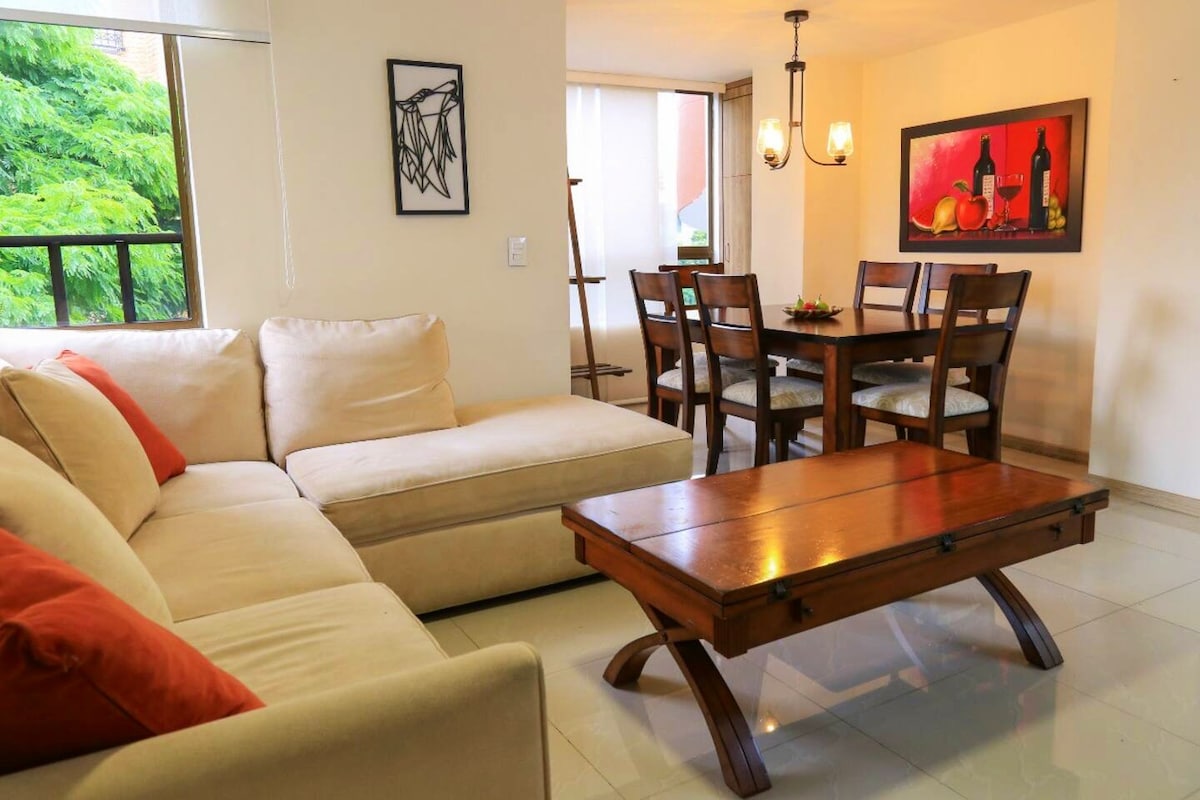 A bright living area features a light-colored sectional sofa adorned with orange pillows. A wooden coffee table is positioned in front of the sofa, while a dining table with six chairs is visible in the background. Natural light filters through large windows, enhancing the spacious ambiance.